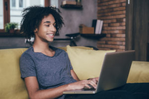 Веселый молодой африканский человек работает на ноутбуке и улыбается, сидя на диване у себя дома