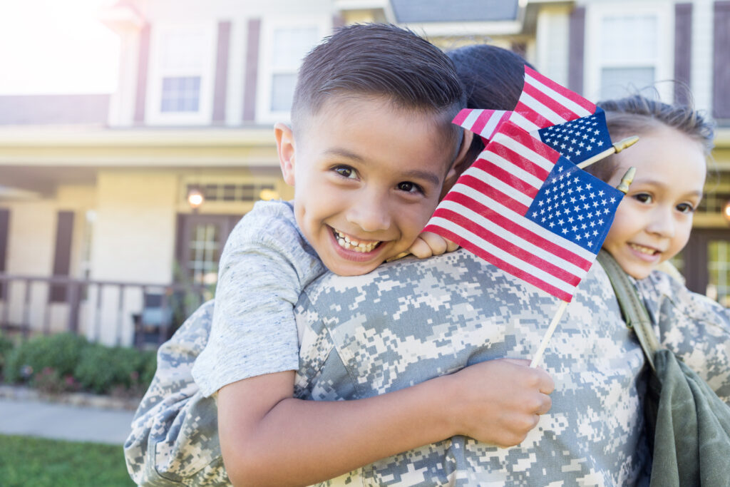 Young boy and girl smiling holding American flags as they hug their mother wearing military camouflage