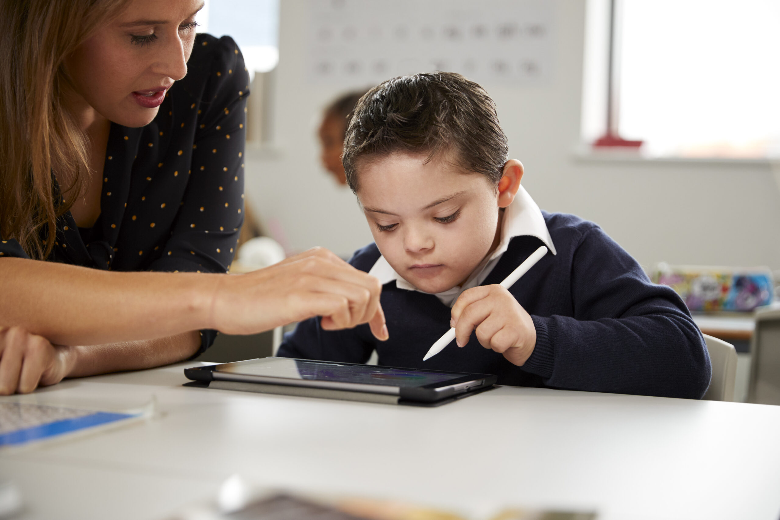 teacher working with a Down syndrome schoolboy sitting at desk using a tablet computer