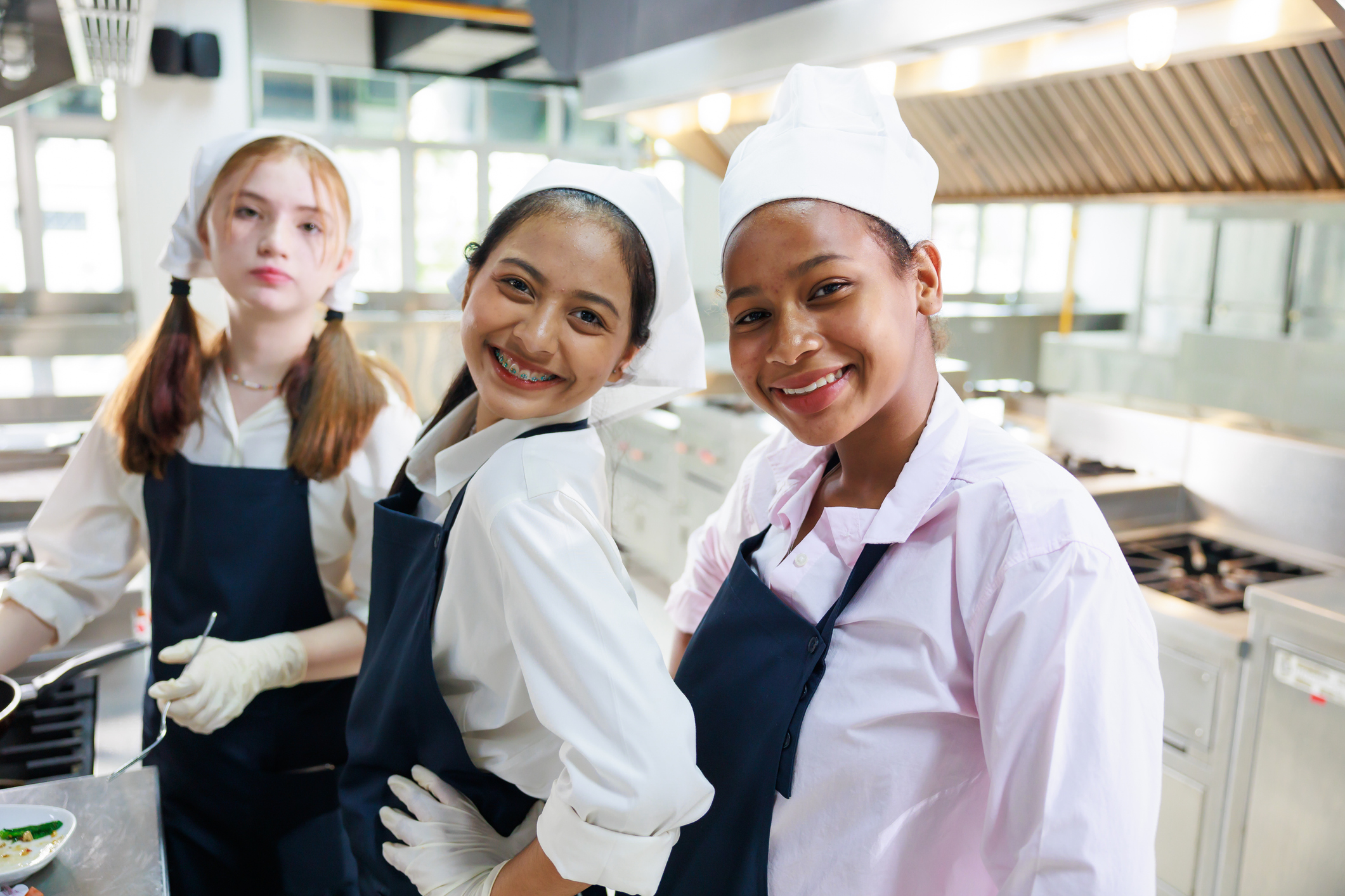 3 teen girls wearing chef's hats and aprons in a kitchen