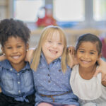 Three little girls of different ethnicities and abilities smile and put their arms around each other in a kindergarten classroom.
